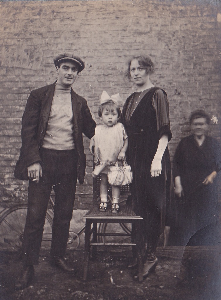 Black and white family photo from many years ago with a gentleman, lady and child who is standing on a chair. Bicycle and older lady in the background.