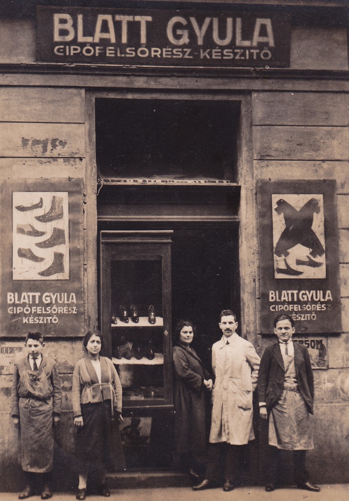 Shoe shop from 1923 with two ladies and three men stood outside. Shows the name of the shop 'Blatt Gyula' on a sign and shiny shoes in the window.