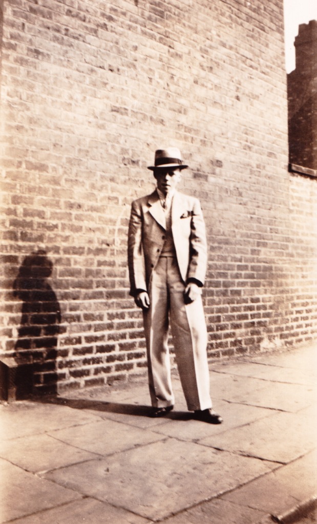 black and white image of a young man in suit and trilby having his portrait taken standing against a brick wall, shadow against the wall.